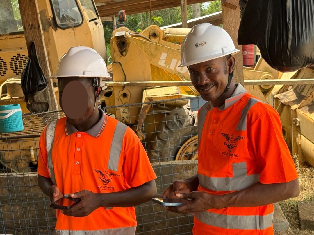 Gold Empire Resources field team in branded orange safety gear and helmets at an active mining site with earth-moving equipment